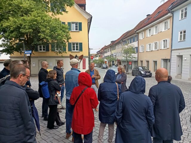 Mehrere Menschen stehen vor Gebäuden und hören dem Vortragenden zu
