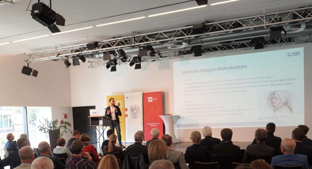 Mann steht auf einer Bühne vor dem Publikum und erzählt. Im Hintergrund ist eine Präsentation auf der Leinwand zu sehen.