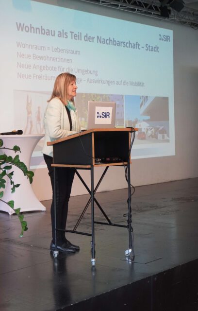 Frau steht auf einer Bühne vor dem Publikum und erzählt. Im Hintergrund ist eine Präsentation auf der Leinwand zu sehen.