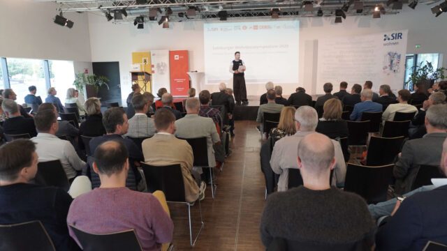 Moderator Wojciech Czaja steht auf der Bühne und begrüßt die Teilnehmenden.