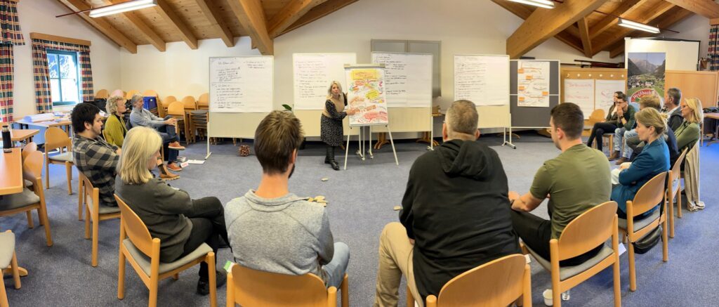 Eine Gruppe Weißbacher Bürger:innen sitzt im Halbkreis, vor einigen Pinnwänden und Flipcharts. Eine Moderatorin erklärt mithilfe eines Flipcharts