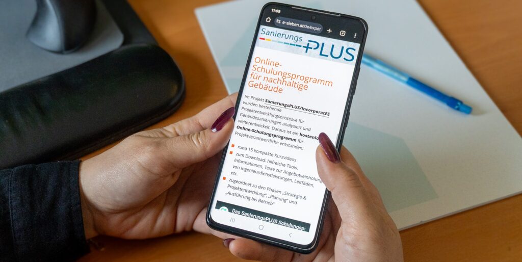 Zwei Hände halten ein Smartphone, auf dem eine Website zum Projekt SanierungsPLUS zu sehen ist