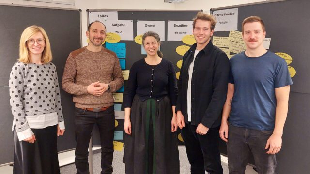 Drei Männer und zwei Frauen stehen vor Pinnwänden.