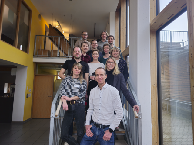 12 Personen stehen auf einer Treppe in einem Gebäude mit viel Glas und Holz