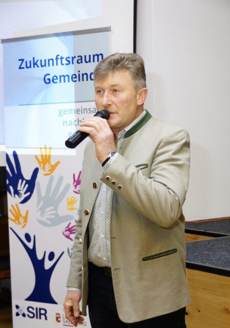 Ein Mann spricht in ein Mikrofon. Im Hintergrund sieht man ein Rollup mit der Aufschrift 