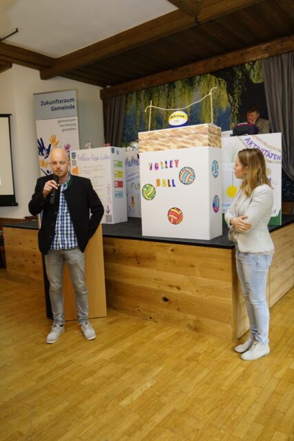 Ein Mann und eine Frau stehen vor selbstgebastelten Kisten. Auf den Kisten stehen verschiedene Projektnamen wie 