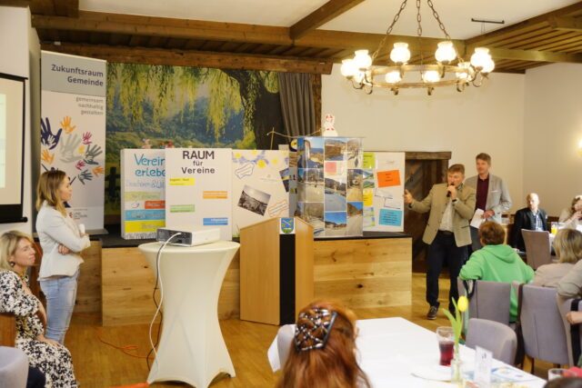 Ein Mann mit Mikrofon präsentiert einem Publikum verschiedene Kisten auf einer Bühne. Auf den Kisten stehen Namen von Projekten aus der Gemeinde Krispl-Gaißau, wie zum Beispiel 