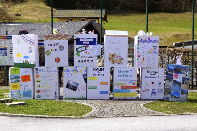 Viele bunte Kisten stehen im Freien. Auf den Kisten stehen die Namen von Projekten aus der Gemeinde Krispl-Gaißau. Zum Beispiel die Kindersachenbörse oder das Gschäftl.