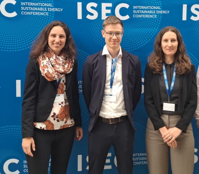 Zwei Männer und zwei Frauen stehen vor einer Wand mit ISEC-Logo