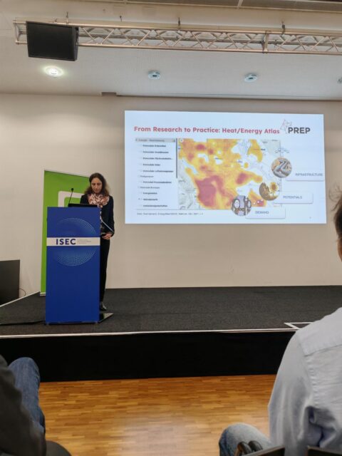 Eine Frau steht auf einer Bühne mit einer Powerpoint-Folie im Hintergrund. Auf der Folie steht 
