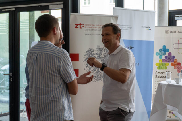 Drei Männer unterhalten sich miteinander. Im Hintergrund stehen Rollupts von der ZT Kammer, klimaaktiv und Smart City Salzburg