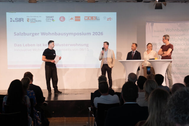 Ein Moderator und vier politische Gäste stehen auf einer Bühne. Im Hintergrund steht auf einer Powerpoint-Folie 