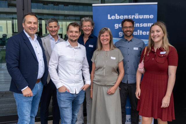Sieben Personen stehen vor einem Rollup mit der Aufschrift 