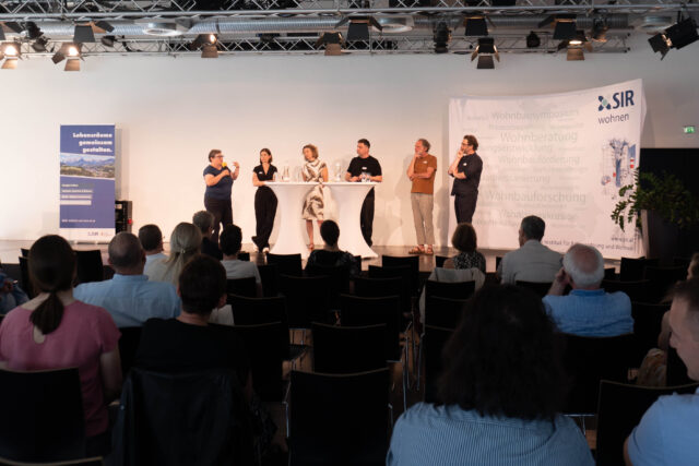 Drei Frauen und drei Männer stehen auf der Bühne des Wohnbausymposiums. Eine Frau hält ein Mikrofon und spricht. Die anderen Personen hören zu