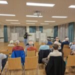 Eine Frau erklärt ein Plakat. Davor sitzt ein Publikum und hört zu.