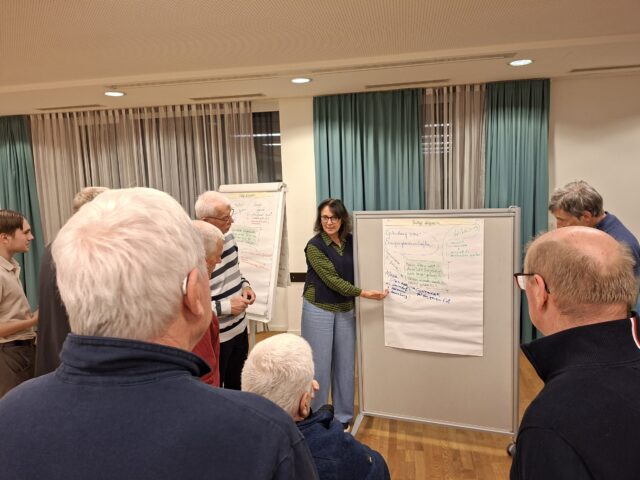 Eine Frau steht vor einer Pinnwand mit einem Plakat und zeigt auf das Plakat.