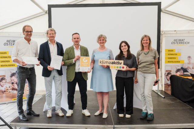 Sechs Personen stehen auf einer Bühne und halten ein Holzschild mit dem klimaaktiv-Logo.